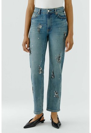 Vrouw die blauwe denim jeans draagt met bloemrijke paillettenborduurwerk, witte top en zwarte puntige slingback schoenen, staand op een effen achtergrond.