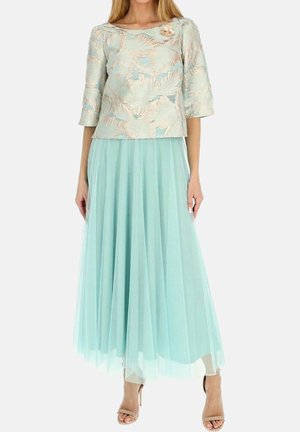 Kobieta ubrana w bluzkę w jasno-niebieskie kwiatowe hafty z rękawami o długości trzy czwarte oraz zwiewną maxi spódnicę z tiulu, w połączeniu z beżowymi szpilkami.