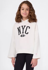 Sudadera blanca con "NYC 5192" impreso en negro, presenta una silueta relajada, mangas anchas y un detalle de logo en el puño. Usada sobre pantalones negros.