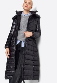 Lang sort pufferjakke, quilted design, blankt materiale, med hætte og lynlås. Båret over en grå sweater og en blåstribet skjorte.