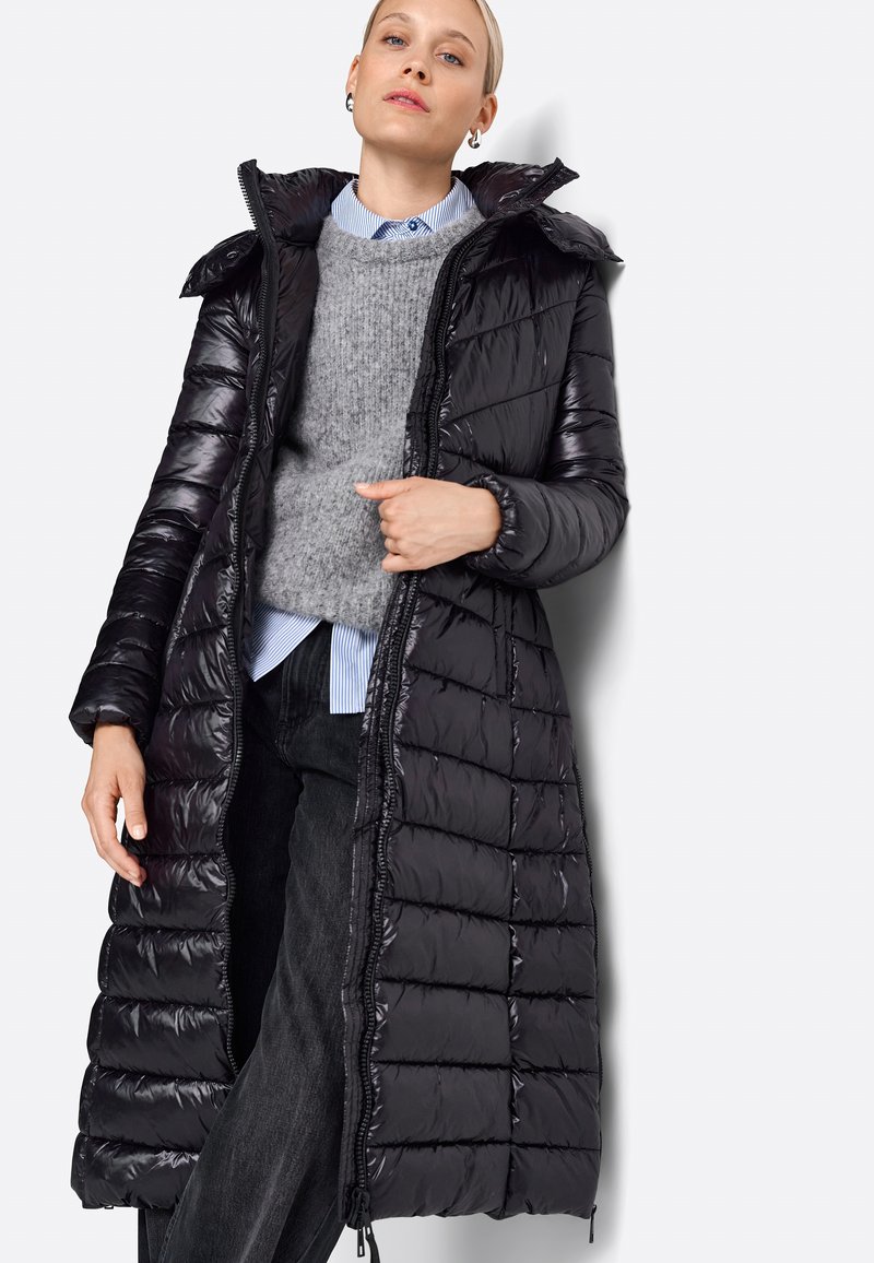 Lang sort pufferjakke, quilted design, blankt materiale, med hætte og lynlås. Båret over en grå sweater og en blåstribet skjorte.
