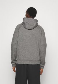 Homme aux cheveux courts portant un sweat à capuche gris et un pantalon noir, debout de dos sur un fond blanc uni.