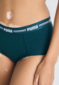 Turkizne bombaške hlačke z Puma pasom, ki ima bele proge. V bližnjem načinu prikazuje logotip pasu in gladko teksturo.