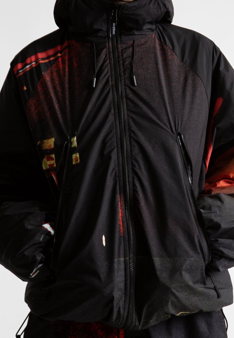 Veste à capuche noire zippée avec des motifs abstraits rouges et jaunes et des poches latérales zippées, portée les mains dans les poches.