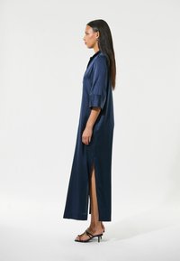 Abito lungo in satin blu navy con maniche corte e spacchi laterali, dotato di colletto e vestibilità rilassata, abbinato a tacchi neri con cinturini.