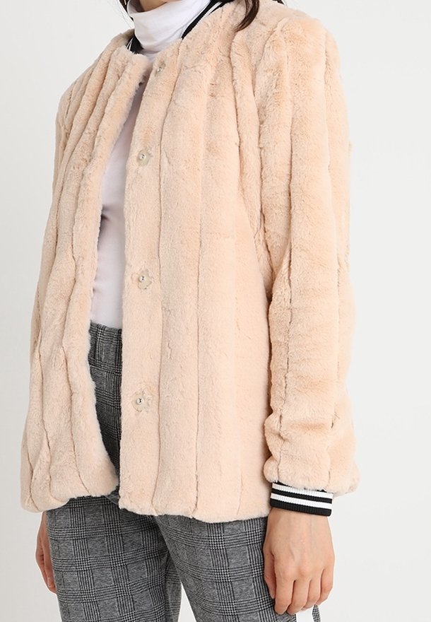 Femme portant une veste boutonnée en peluche beige clair par-dessus un col roulé blanc et un pantalon à carreaux gris, sur un fond uni.