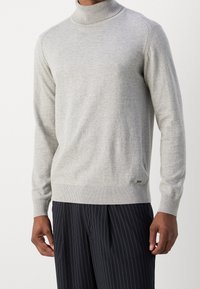 Grå rullekrave-trøje med ribbet krave og kant, lavet af blød tekstur. Har et diskret logodetalje nederst på venstre side.