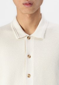 Polo en tricot couleur crème avec un tissu texturé, fermeture à trois boutons et un col doux. Présente des boutons ronds marron clair.