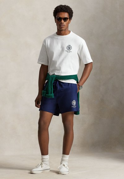 Hvit bomulls-t-skjorte med logo, marineblå shorts, grønn genser knyttet rundt livet, hvite joggesko og solbriller; avslappet passform og uformell stil.