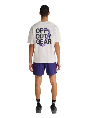 Mand iført hvid t-shirt med sort "OFF DUTY GEAR" tekst og lilla karabinhage grafik, navy shorts, sorte sokker og grå sko, vendt væk.