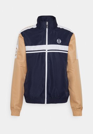 Marineblå træningsjakke med hvide vandrette striber, beige ærmer, elastiske manchetter, fuld lynlås foran og lille hvid logo på brystet.