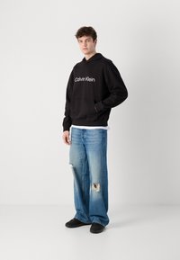 Juoda gobtuvinė palaidinė su "Calvin Klein" logotipu, priekinė kišenė, dėvima su nunešiotais šviesiai mėlynais plačiomis kelnių ir juodais batais.