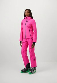 Γυναίκα ντυμένη με φωτεινή ροζ μπουφάν και παντελόνι σκι, φορώντας μαύρα και πράσινα μποτάκια σκι, στέκεται μπροστά σε απλό λευκό φόντο.