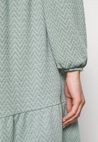 Robe texturée vert clair avec un motif en zigzag, manches longues bouffantes et ourlet froncé, présentant une silhouette incurv ée.