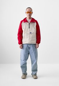 Roter und beige Kapuzenpullover mit einer Fronttasche, schwarzem Reißverschluss und elastischen Bündchen. Hellblaue weite Jeans und beige Schuhe.