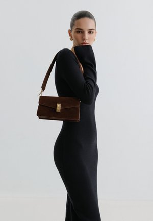 Vrouw in een nauwsluitende zwarte jurk met lange mouwen poseert met een bruine leren schoudertas en gouden oorringen tegen een effen lichte achtergrond.