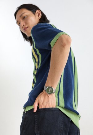 Polo en tricot rayé bleu et vert à manches courtes, porté avec un jeans en denim noir et une montre en argent avec un cadran vert.