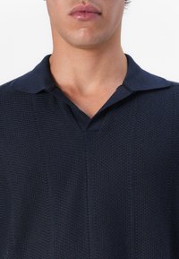 Homme portant un polo en maille texturée bleu marine foncé avec col, visible du cou jusqu'au milieu de la poitrine sur fond blanc.