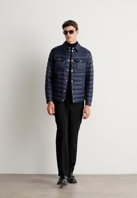 Mornarska puhovka z gumbom spredaj, dvema žepoma na prsih in rebrastim ovratnikom, v kombinaciji z črnimi hlačami in svetlečimi črnimi čevlji.