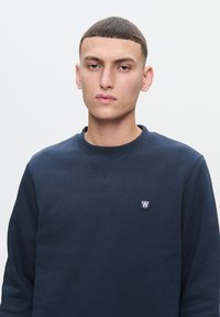 Navy blue sweatshirt with a round neckline and ribbed cuffs, featuring a small, circular logo on the chest. Smooth texture throughout.