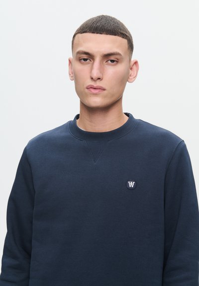 Sweatshirt bleu marine avec un col rond et des poignets côtelés, arborant un petit logo circulaire sur la poitrine. Texture lisse sur l'ensemble.