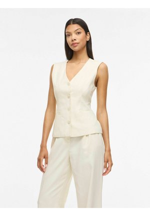 Kvinde iført ærmeløs cremefarvet knapvest og matchende vidde bukser, stående mod en ensfarvet hvid baggrund.