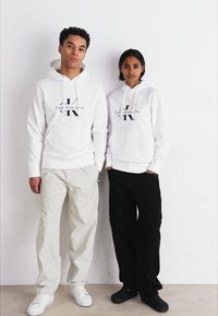 Dva pojedinca nose bijele dukserice s crnim Calvin Klein logotipom. Jedan par ima bež hlače, a drugi crne hlače. Obojica nose tenisice.