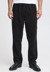 Pantaloni di velluto a coste neri con vita regolabile con coulisse, caratterizzati da una texture a coste verticali e gambe dritte, abbinati a scarpe di pelle nere.