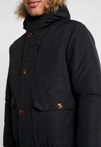 Sort isoleret parka med pelskantet hætte, har brune knapper, lynlåslomme og sidelommer. Glat stof og afslappet pasform.