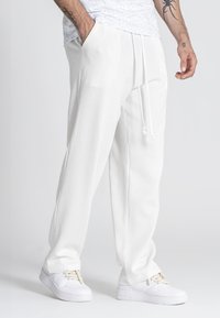 Pantalones jogger blancos, texturizados, con cinturilla elástica, cordones y bolsillos laterales, combinados con zapatillas blancas.