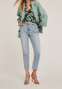 Světlý mátový oversize kabát, černá květinová blůza, světlezelené rovné džíny a sandály na vysokém podpatku s dekoračním akcentem.