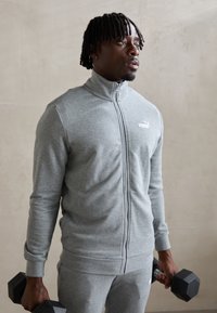 Veste de sport zippée grise avec manches longues et col haut, arborant un petit logo blanc. Le modèle tient des haltères noirs.