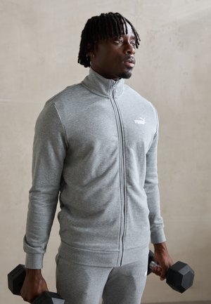 Gray zip-up athletic jacket with long sleeves and a high collar, featuring a small white logo. The model holds black dumbbells.