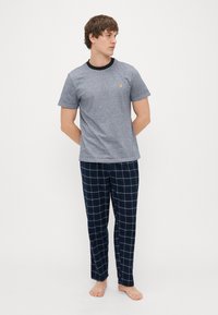 T-shirt gris à manches courtes avec col noir et petit logo jaune, associé à un pantalon de pyjama à carreaux bleu marine avec des lignes blanches.