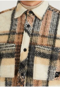 Koszula polarowa w beżowym, brązowym i szarym wzorze w kratę. Posiada zapięcie na guziki z przodu, dwie kieszenie na piersi oraz klasyczny krój kołnierzyka.