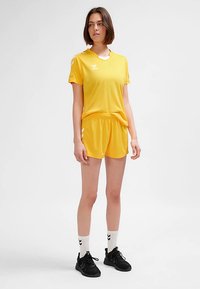 Uniforme de sport jaune à manches courtes, col rond, et short assorti. Présente un logo et des accents blancs contrastants. Porté avec des baskets noires.