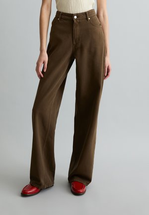 Person trägt hoch taillierte, locker sitzende braune Hose mit Fronttaschen und roten Schuhen, steht vor einem einfarbigen grauen Hintergrund.