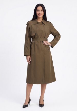 Frau mit langen dunklen Haaren, die einen olivgrünen Trenchcoat mit Gürtel und schwarze High Heels trägt, steht vor einem weißen Hintergrund.