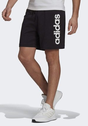Mężczyzna w czarnych spodenkach Adidas z białym logo z boku i białych butach sportowych, stoi z jedną nogą skrzyżowaną z przodu.