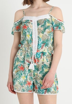 Frau trägt einen floralen Schulterfreien Jumpsuit mit Spitzenbesatz und einem weißen Häkel-Detail mit Quasten in der vorderen Mitte.