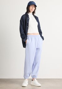 Heledad sinine dressipüksid elastsete varrukatega, kombineerituna valge kõrge kaelusega T-särgi ja tumedast denimist nööpidega särgiga, viimistledes välimust valgete spordijalatsitega.