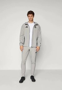 Jachetă sportivă de culoare gri deschis, cu glugă și accente negre, asociată cu pantaloni ajustați de aceeași nuanță. Prezintă un logo brodat și fermoare.