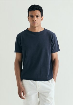Bărbat tânăr cu păr scurt și creț, purtând un tricou bleumarin cu mâneci scurte și pantaloni albi, stând în fața unui fundal simplu și deschis la culoare.