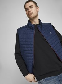 Navy quilted vest with a stand collar, featuring a contrasting zipper and a subtle logo. Worn over a black long-sleeve shirt.