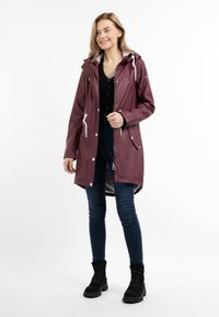 Jachetă maro waterproof cu glugă, închidere cu fermoar și capse, șnururi albe, buzunare laterale și un tiv ușor mai lung în spate.