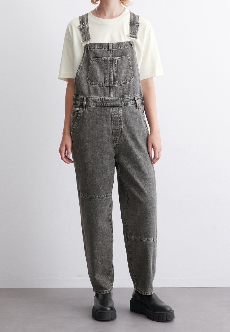Personne portant une salopette en jean noir délavé, un t-shirt blanc à manches courtes et des bottes noires à semelle épaisse, debout contre un mur blanc.