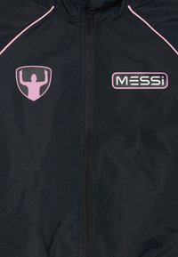 Svart jacka med central dragkedja, rosa piping på axlarna, en rosa silhuettlogotyp med uppsträckta armar på vänster bröst och "MESSI"-text på höger bröst.