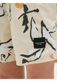 Shorts de bain beige avec un motif floral vert et orange. Présente une étiquette noire avec "JACK & JONES EST. 1990" près de l'ourlet.