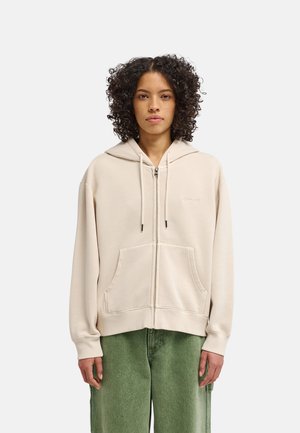 Persona con capelli ricci indossa una felpa beige con zip e tasche frontali e pantaloni verdi, in piedi contro uno sfondo chiaro e semplice, rivolta in avanti.
