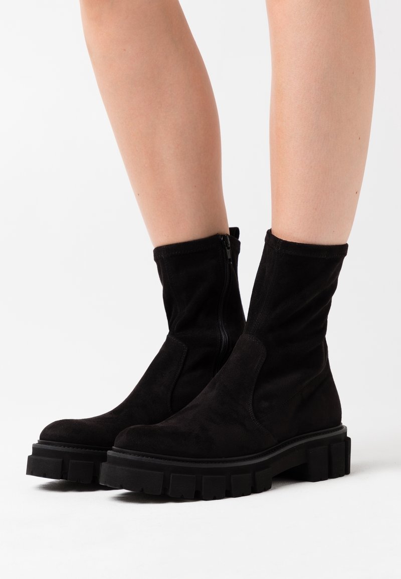Jambes portant des bottines en daim noir avec des semelles en caoutchouc épaisses et robustes et des fermetures éclair latérales sur fond blanc.